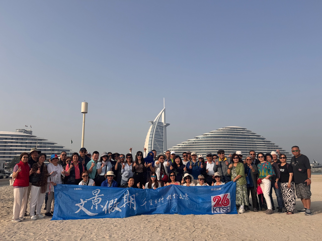 A large group of people stands on a sandy beach holding a blue banner with Chinese text, with the Burj Al Arab and a large hotel visible behind them.